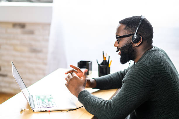 African-American guy with headset using laptop Concentrated young guy in smart casual wear and stylish glasses is using headset and laptop for online communication, supporting, selling. A black guy sits at the office desk, side view sports training stock pictures, royalty-free photos & images