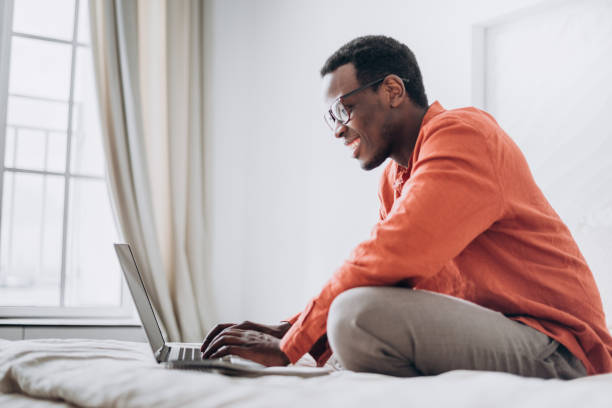 African-American man freelancer works on laptop on bed stock photo