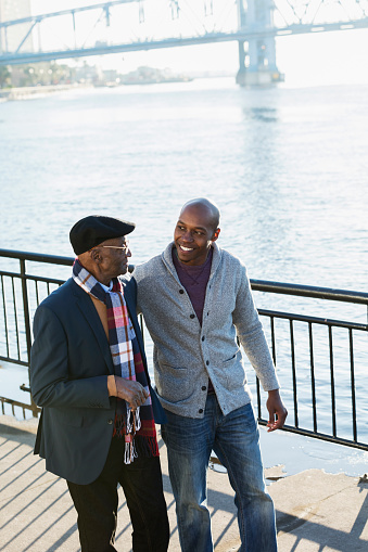 A mid adult African-American man in his 30s standing on a city waterfront with his grandfather, a senior man in his 70s, smiling and talking. It is a sunny autumn day.