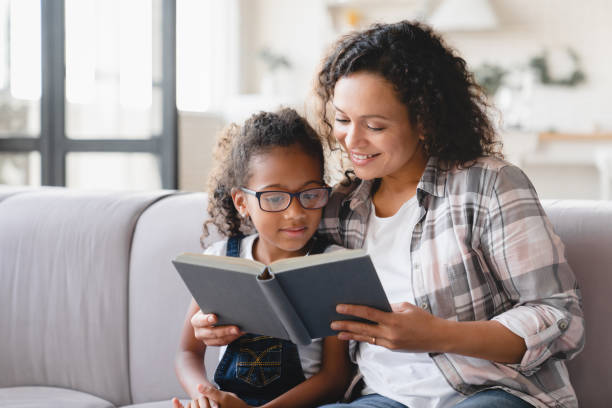 African-american mother mom reading fairy tales book novel with daughter girl, learning together, helping assisting with homework for school. Female tutor teaches schoolgirl at home. Homeschooling stock photo