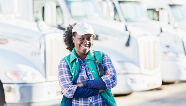 African-American woman standing in front of semi-trucks A mid adult African-American woman in her 30s, a female truck driver standing in front of a fleet of semi-trucks or tractor-trailers parked in a row. Her arms are crossed and she is looking at the camera, smiling confidently. truck driver stock pictures, royalty-free photos & images