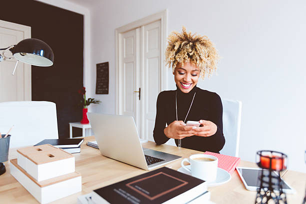 Afro american young woman in a home office Beautiful afro american young woman sitting at the desk in a home office and using a smart phone. contented emotion stock pictures, royalty-free photos & images