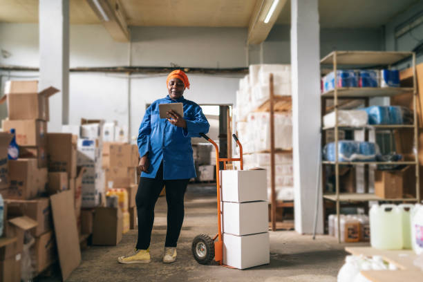 Afro female warehouse worker using a hand truck and checking delivery on digital tablet stock photo