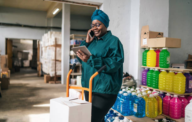 Afro female warehouse worker using a hand truck and talking on a smart phone in distribution warehouse stock photo
