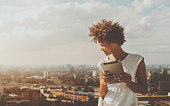 Cheerful young black curly female leaning on railing of balcony of skyscraper, holding digital tablet and looking down on city from high above; with copy space place for text, your message or advert