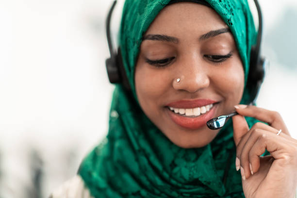 Afro Muslim female with green hijab scarf customer representative business woman with phone headset helping and supporting online with customer in modern call centre stock photo