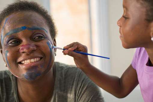 An afro-american  girl is painting her mother's face with tempera at living room table