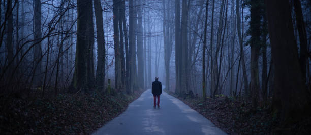 Aged man alone in misty forest, standing lonely and abandoned on an empty road in a gloomy atmospheric mood stock photo