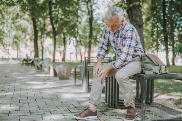 Aged man having sudden ache in his knee stock photo Serious mature man on the bench in the park putting hands on the knee knee stock pictures, royalty-free photos & images