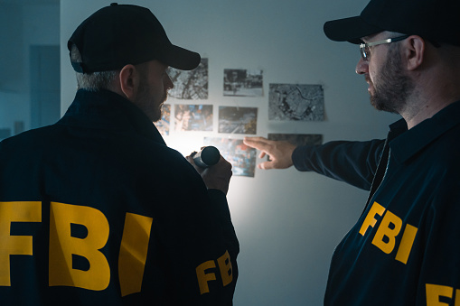 Two FBI agents at work. Looking at photos on the wall at victim's apartment, we see them from the waist up from the back.