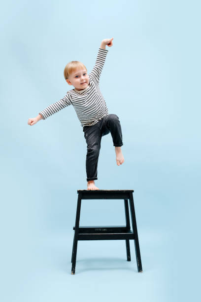 Agile blond boy balancing on one foot on a stepping stool. Over blue background stock photo