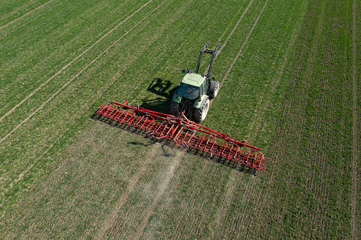 Green, modern, tractor with a red harrow working in field in spring, aerial view.