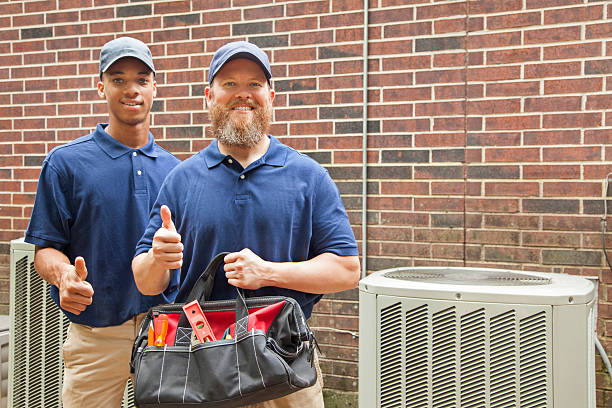 Air conditioner repairmen work on home unit.  Tool bag. Multi-ethnic team of men repairing a home's air conditioner unit outdoors. They have completed the repairs.  They both wear blue uniforms and man in foreground holds a tool bag.  Thumbs up for a job well done! technician stock pictures, royalty-free photos & images