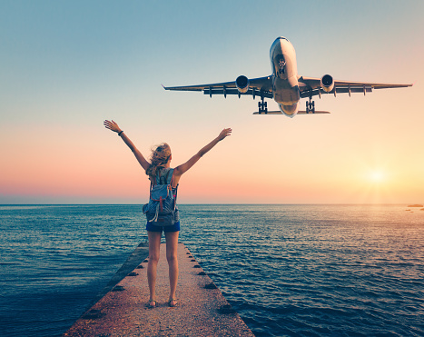 Airplane and woman at sunset. Summer landscape with girl standing on the sea pier with raised up arms and flying passenger airplane. Woman and landing commercial plane in the dusk.Lifestyle and travel