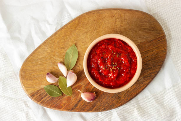 Ajvar in a bowl, garlic and spicy leaves on a wooden board. Serbian traditional food. Ajvar, balkan traditional sauce in a bowl, garlic and spicy leaves on a wooden board. Serbian traditional food. Top view. chutney stock pictures, royalty-free photos & images
