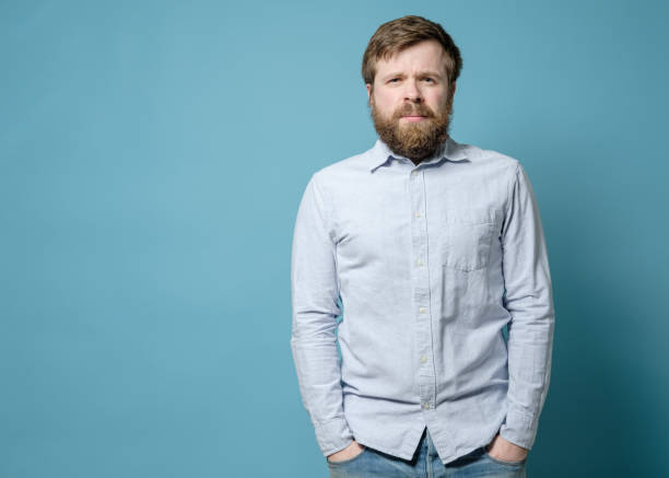 Alarmed Caucasian man with a shaggy beard and an overgrown hairstyle stands with hands in jeans pockets. Copy space. stock photo