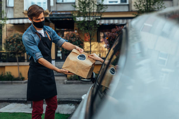ale restaurant worker giving a packed food to a customer in front of a restaurant. Restaurant worker wearing a protective face mask delivering a bag with fresh food to a customer. curbsidepickup stock pictures, royalty-free photos & images