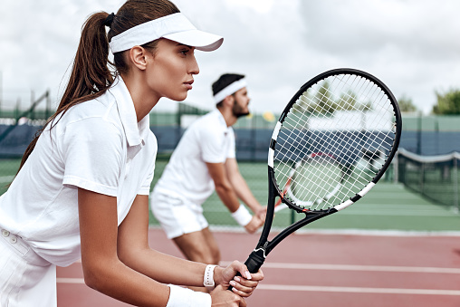 Young woman and man in white sportswear stand on the court with rackets in their hands fully concentrated on the game