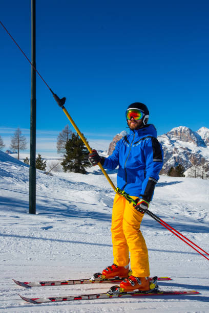 Alpine skier with T-bar lift Alpine skier with T-bar lift, blue and yellow clothes on slope with mountains in the background at Cortina d'Ampezzo Faloria skiing resort area Dolomiti Italy t bar ski lift stock pictures, royalty-free photos & images