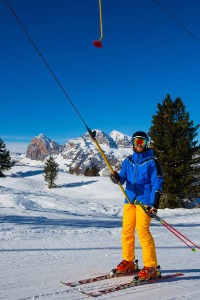 Alpine skier with T-bar lift Alpine skier with T-bar lift, blue and yellow clothes on slope with mountains in the background at Cortina d'Ampezzo Faloria skiing resort area Dolomiti Italy t bar ski lift stock pictures, royalty-free photos & images