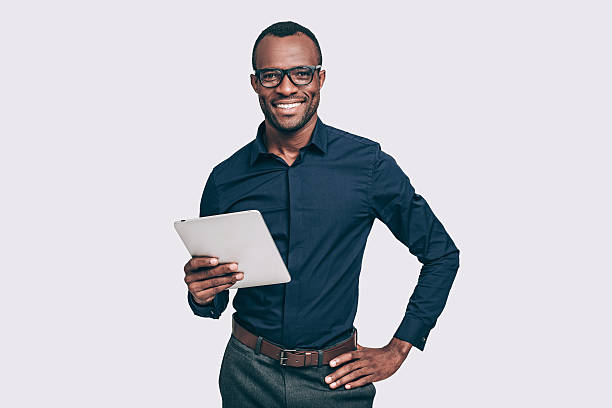 Always ready to help. Handsome young African man holding digital tablet and looking at camera with smile while standing against grey background hand on hip stock pictures, royalty-free photos & images