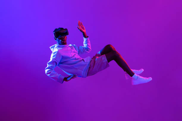 Amazed young African man wearing VR glass headset levitating in the air on futuristic purple cyberpunk neon light background stock photo