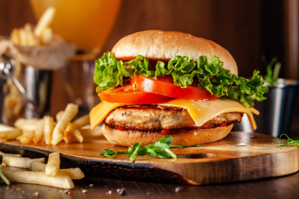 American cuisine concept. Juicy burger with meat patty, tomatoes, cheddar cheese, lettuce and homemade bun. In the background are french fries and a glass of beer. Close up stock photo