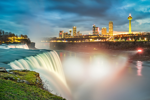 Long exposure photo of the American Falls waterfall in New York State, USA and the skyline of the city of Niagara Falls, Ontario, Canada, in the background, illuminated at twilight blue hour.