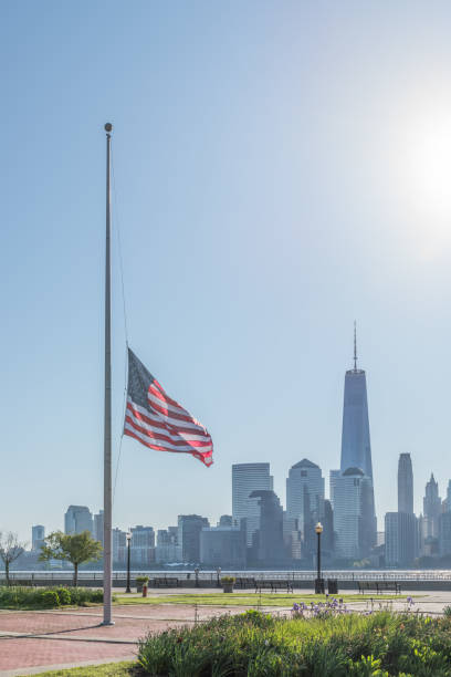 American flag flying at half mast with the New York City skyline in the background. American flag in the Liberty Science Center in Jersey City at half mast, in a vertical photo with the sun rising in the background, casting morning light on the flag. flag at half staff stock pictures, royalty-free photos & images