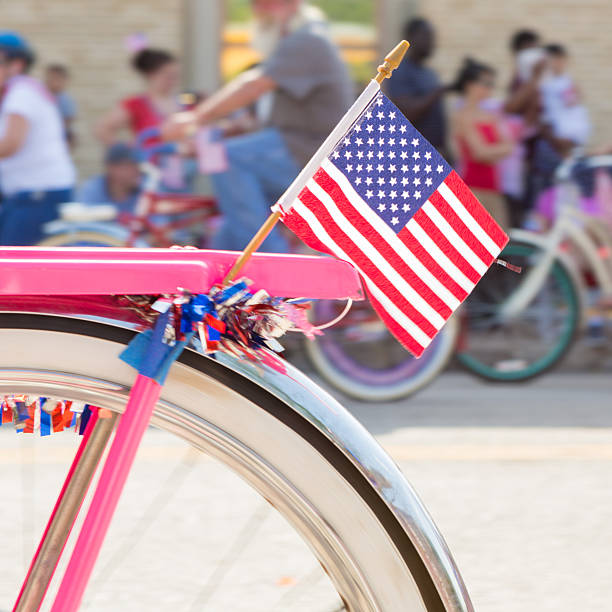 American flag on bicycle during parade on Fourth of July Small American flag is attached to pink vintage bicycle. Bike is decorated for Fourth of July or Independence Day and appears to be riding on street in city during parade. Other people and bicycles can be seen in background. parade 4th of july stock pictures, royalty-free photos & images