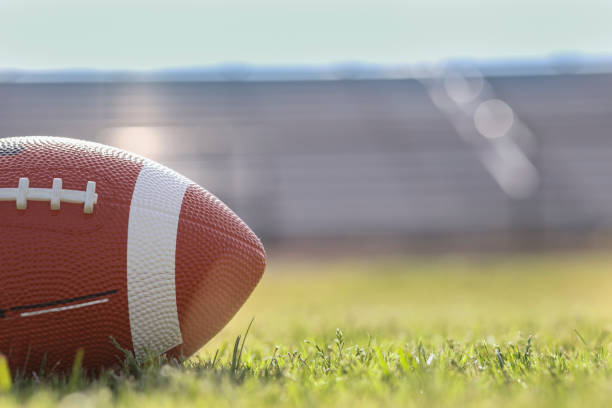 American football on stadium field at school campus. Football on grass stadium on college or high school campus. Bleachers background. No people.  Daytime. football stock pictures, royalty-free photos & images