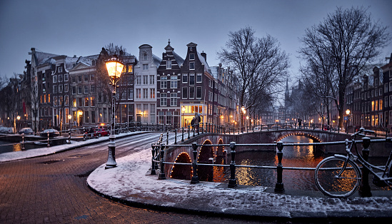 Amsterdam canals and typical canal houses in the old part of the city during a night in winter. The streets are covered in white from the snow, the city lights are on and the sky is grey because of the clouds. You can see some snow falling down.