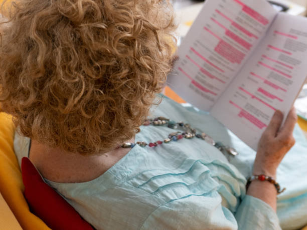 An actress sitting down, learning her lines from a script An actress, with short brown wavy hair, wearing a blue linen dress, sitting on a yellow covered sofa, with a red cushion behind her. She is learning her lines from a film script that is printed on white A4 paper. Her characters part is highlighted with pink highlighter pen. She is wearing a coloured beaded necklace and bracelet. The view is taken from behind her with the script out of focus. film script stock pictures, royalty-free photos & images