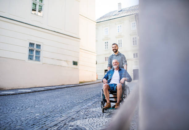 an adult son with senior father in wheelchair on a walk in town. - wheelchair street happy imagens e fotografias de stock