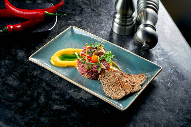 An appetizer before the main course - beef steak tartare served with croutons, cappers, yolk, pickles in a blue plate on a marble background. Restaurant food. Raw meat stock photo