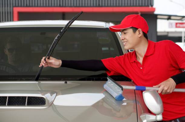 An Asian gas station worker raises the windshield wipers to clean the car windshield. A caucasian driver with sunglass raise thumbs up to express his appreciation from the inside of the car. stock photo