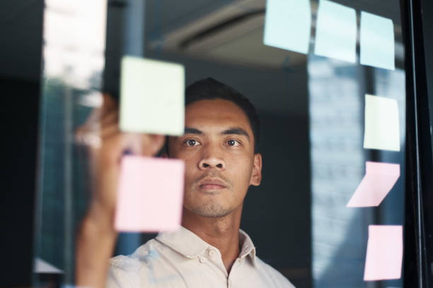 An Asian man brainstorms with sticky notes as seen through a window stock photo