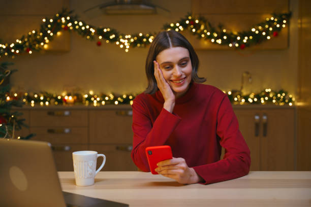 An attractive Christmas girl with a bob haircut in a red sweater is smiling looking in her red phone, sitting at a table on her kitchen with Christmas lights stock photo
