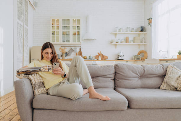 An attractive woman with a bionic prosthetic arm sits at home on the couch with a phone and surfs the Internet on a sunny day stock photo