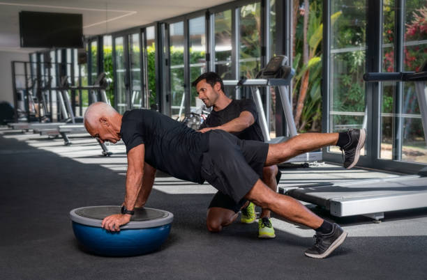 An elderly Caucasian man, in the gym, with a personal fitness trainer, in rehabilitation, doing exercises for balance on a gymnastic ball. stock photo