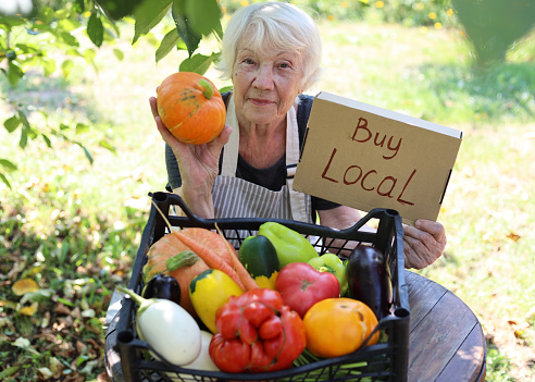 An elderly gray-haired woman sells fresh seasonal vegetables at a local farmers' market. Local business support. Buy local non-GMO agricultural products.