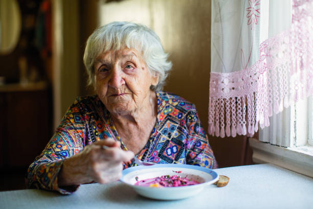 An elderly woman eating soup sitting at a table in the house. stock photo