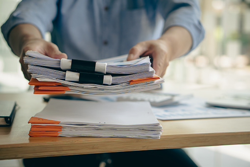 An employee sits at a table full of paper. stamp on the pile of unfinished documents A young account manager's secretary works in the office among piles of paperwork on the table.