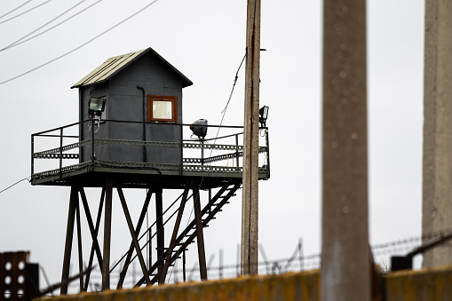 An old gray observation tower made of sheet metal with lantern spotlights and a window shot in the military industrial zone in Russia against the white cloudy sky of pillars and a concrete fence.