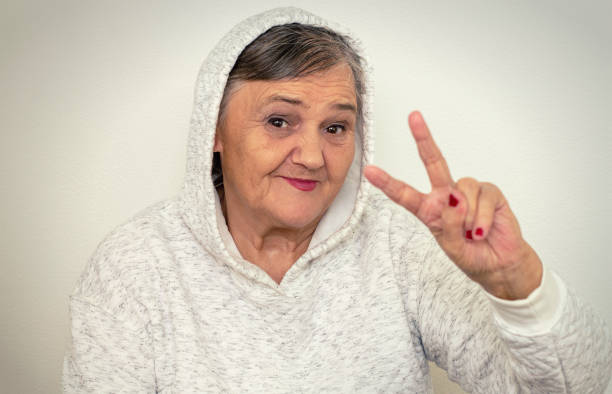 An older hipster woman. Nice shot of a gangster grandma. The old woman dressed in the style of hip-hop, her mood is elated.The old woman shows a peace sign. stock photo