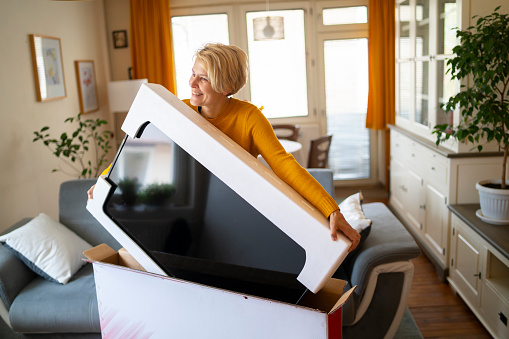A female online shopper takes delivery of her new television and lifts it carefully from its protective packaging inside her modern home