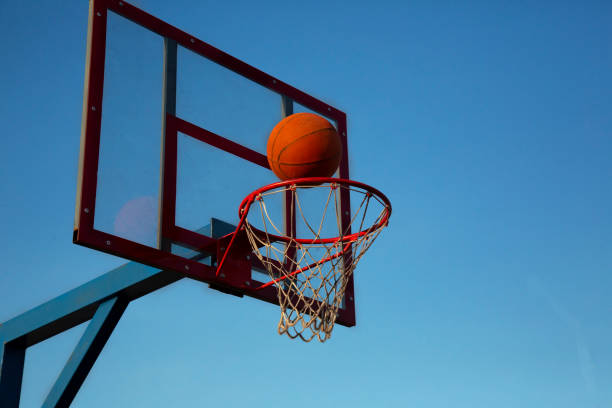 An orange basketball is flying into the basket on an open area against the sky. stock photo