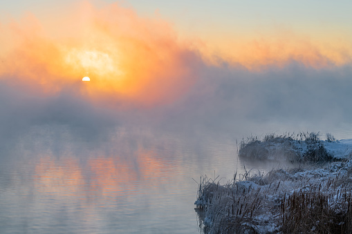 An orange fog spreads over the water and obscures the horizon.  Sunset Sunrise time over the winter river. Reeds stand in the snow near the water.