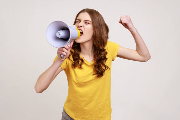 Angry nervous teenager girl with brown hair in yellow t-shirt loudly screaming at megaphone, making announce, protesting, wants to be heard. stock photo