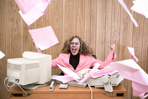 A vintage African American business woman at the office works at an old computer at her desk.  She screams and throws papers into the air in frustration.  1980's - 1990's fashion style.  Wood paneling on wall in the background.